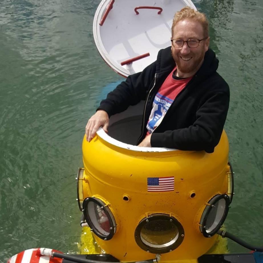 Adam Katz emerging from the hatch of a small yellow personal submarine on the water